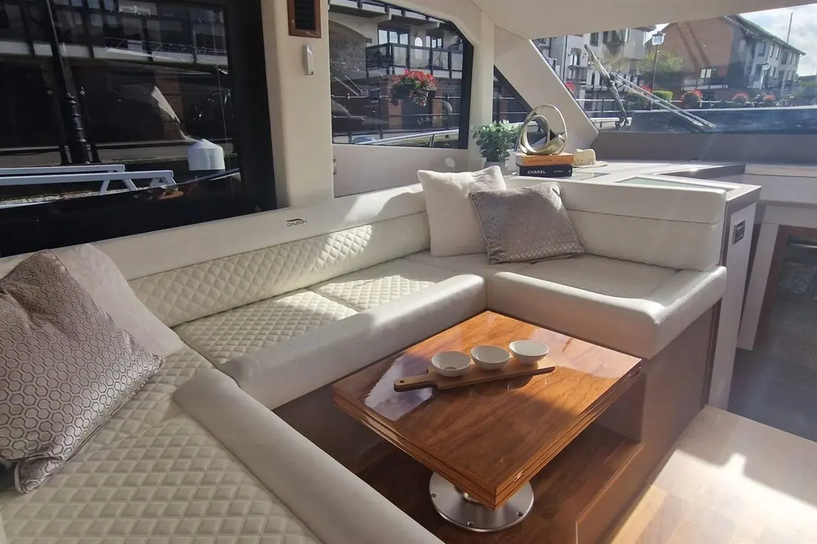 A modern boat interior featuring a white leather sofa, wooden coffee table with bowls, and large windows with a canal view.
